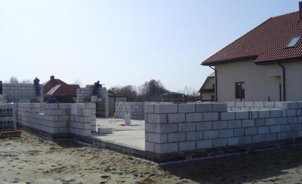 Wznoszenie ścian parteru z bloczków na fundamencie domu jednorodzinnego, widoczni pracownicy w kombinezonach, w tle budynek z czerwoną dachówką.