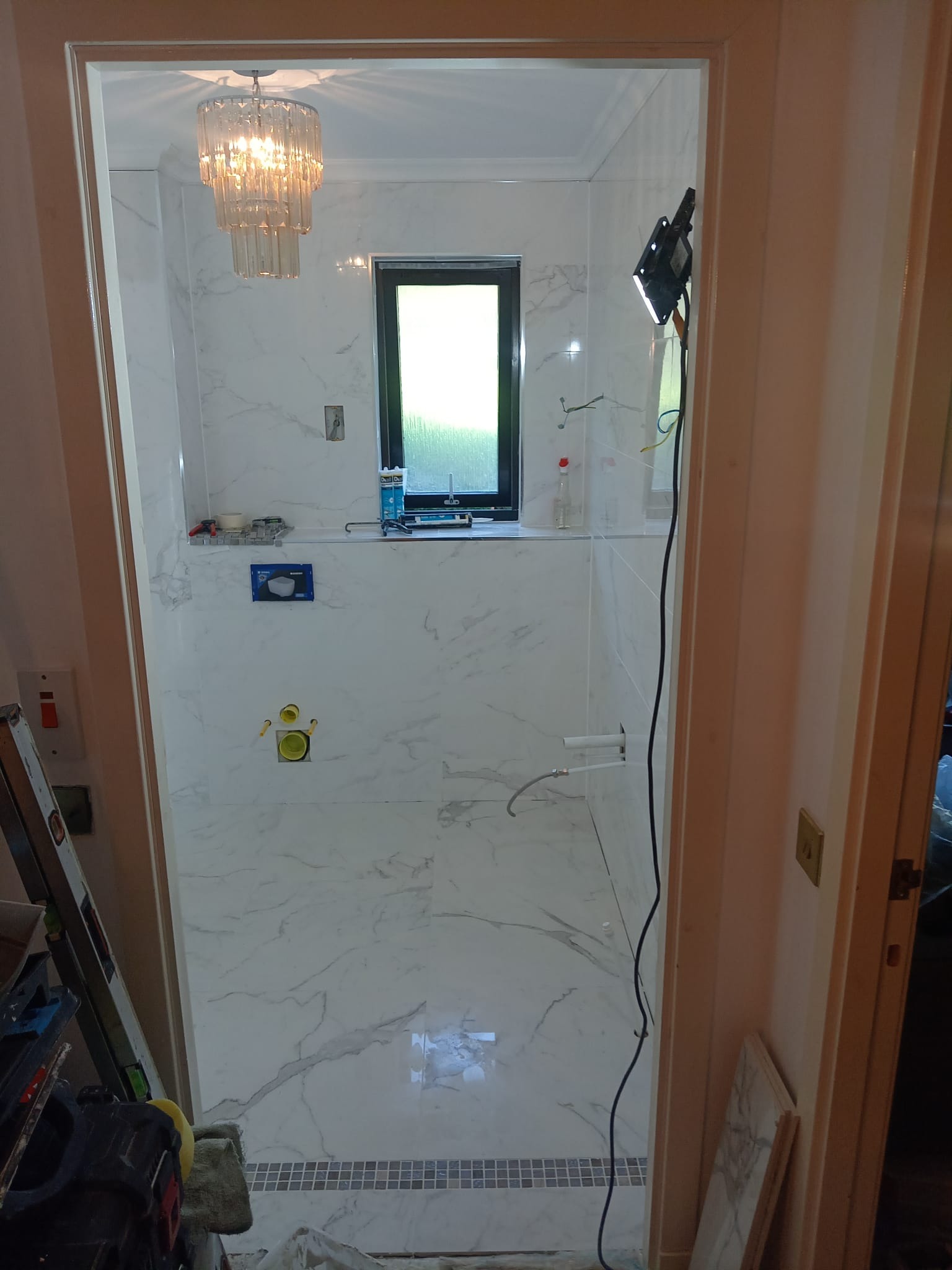 Wnętrze łazienki w trakcie remontu, widoczne białe płytki imitujące marmur na ścianach, małe okno, sufit z oświetleniem oraz elementy instalacji wodnej i kanalizacyjnej przed zabudową, narzędzia...