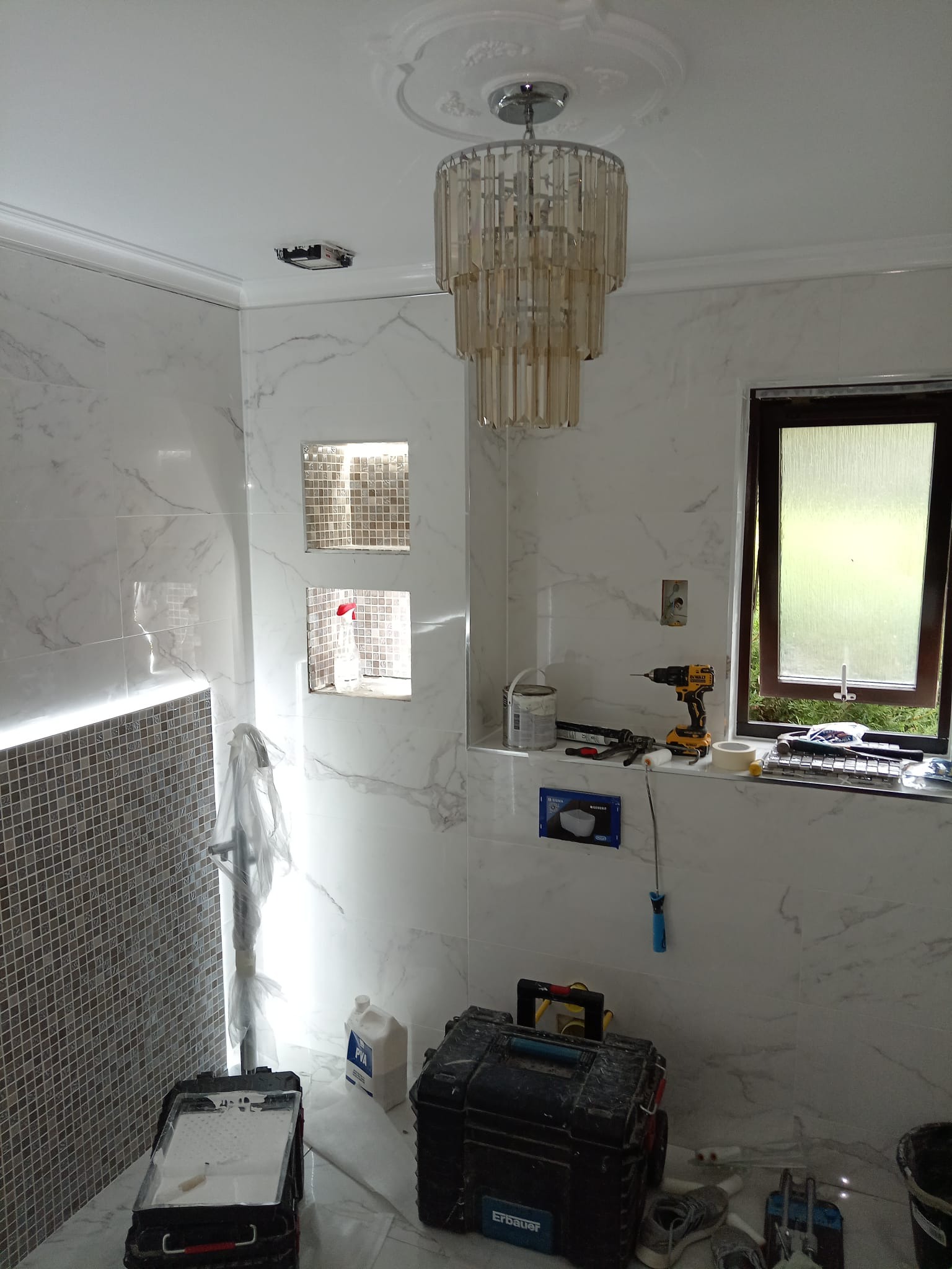 Remont łazienki w trakcie: marmurowe płytki, ozdobny żyrandol, narzędzia i akcesoria malarskie rozłożone na parapecie okiennym i podłodze, wnęki z mozaiką. Widoczne ślady prac remontowych.