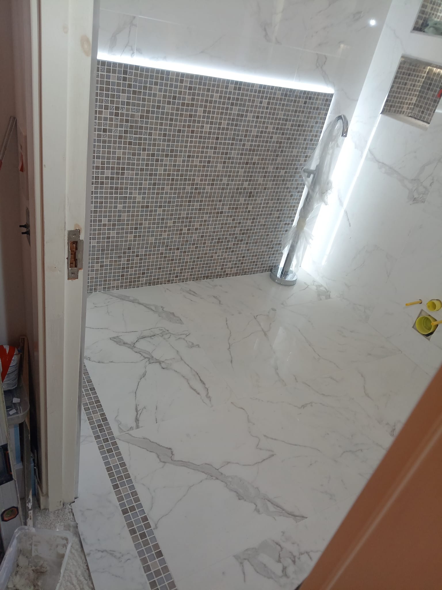 Wnętrze łazienki we Wrocławiu w trakcie remontu, z marmurową podłogą i mozaikową ścianą podświetloną ledami. Widoczna instalacja prysznicowa w folii ochronnej i akcesoria malarskie.