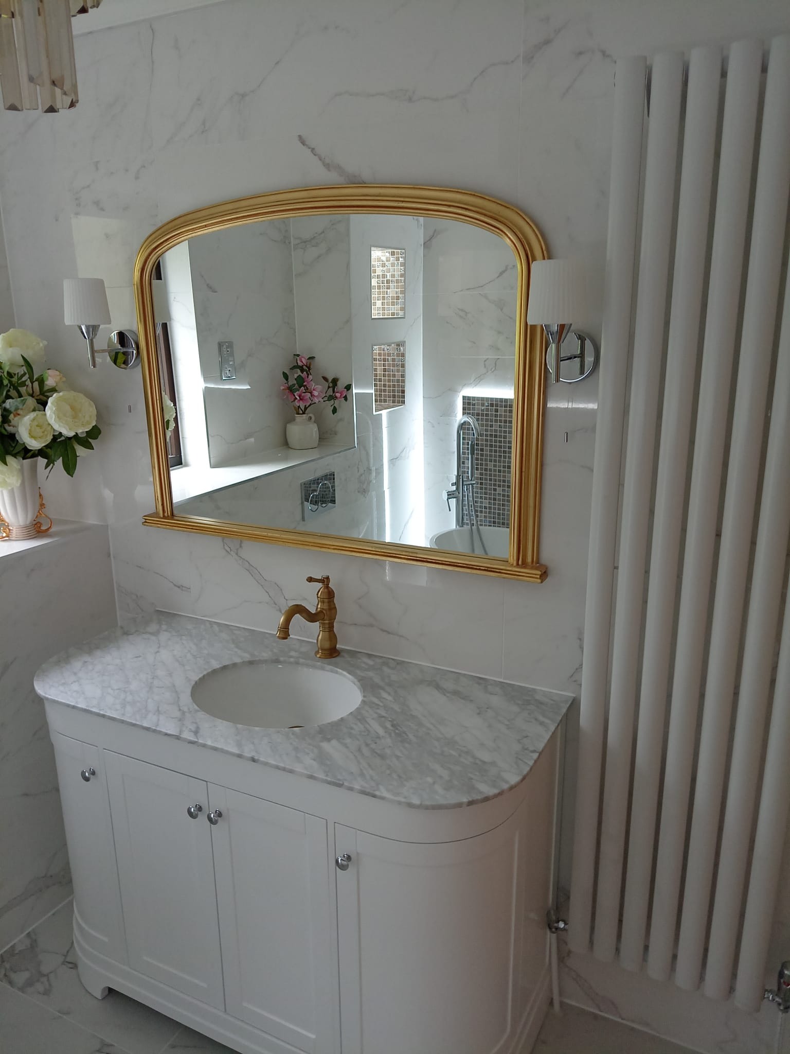 Elegancka łazienka z marmurowymi płytkami, białą szafką pod umywalkę z marmurowym blatem i złotą armaturą, duże lustro w złotej ramie odbija fragment wanny i mozaiki, obok grzejnik żeberkowy.