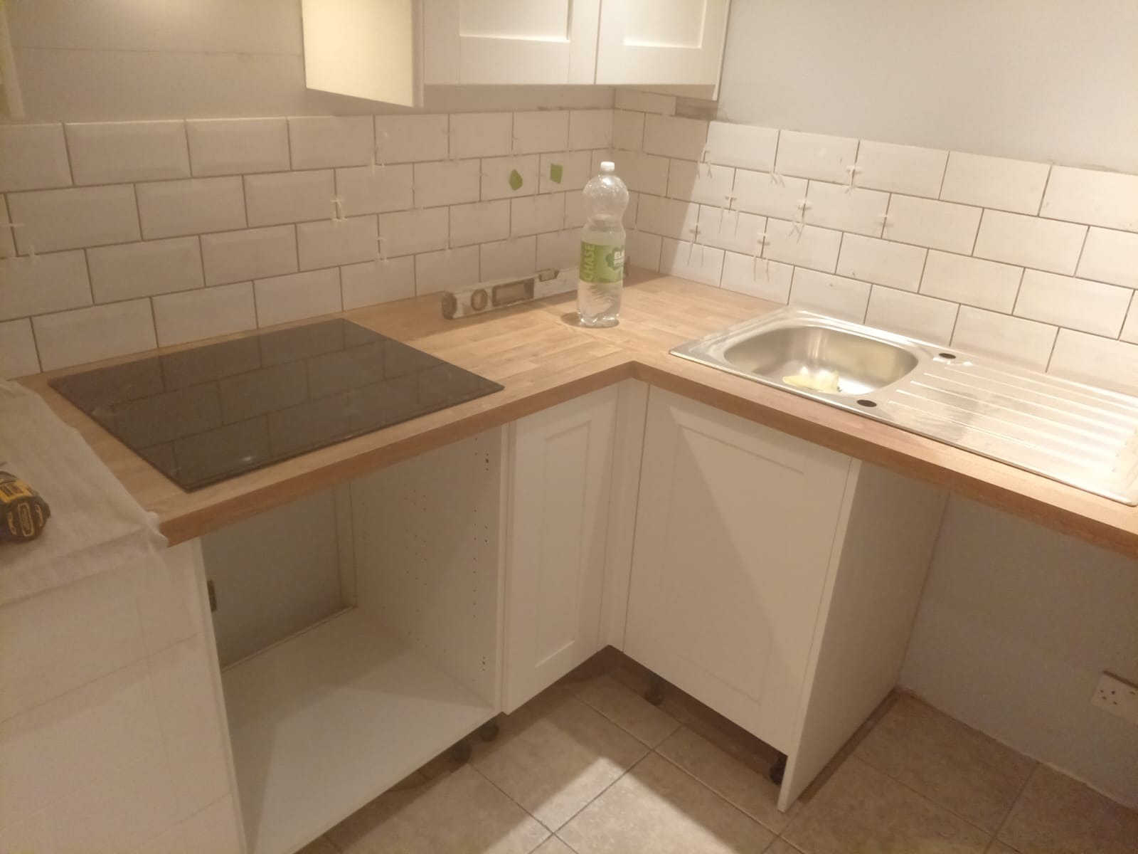 Montaż płytek ściennych w kuchni z widocznymi krzyżykami dystansowymi, drewnianym blatem, zlewem i płytą indukcyjną, butelką wody i poziomicą.