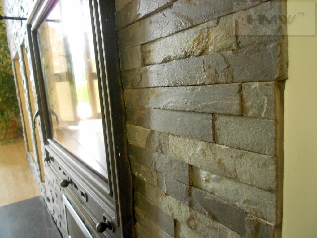 Fragment ściany wykończonej szarą, warstwową okładziną kamienną, przylegającej do czarnej obudowy wkładu kominkowego z widoczną szybą i uchwytami.