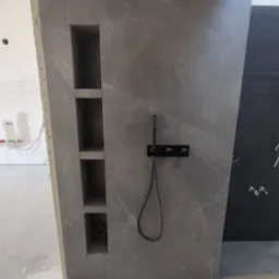 Ściana prysznicowa wyłożona szarą płytką imitującą marmur z wnękami na kosmetyki i czarną armaturą, widoczne niedokończone prace w tle.