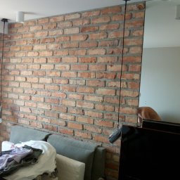 ERKAM,GLAZURNICTWO - Ściana z czerwonej cegły z szarą fugą, fragmentarycznie widoczny szary narożnik sofy i stos ubrań, w tle odbicie w lustrze