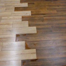 ERKAM,GLAZURNICTWO - Podłoga wyłożona panelami w dwóch odcieniach drewna: jasnym i ciemnym, z widocznymi kroplami wody na powierzchni.