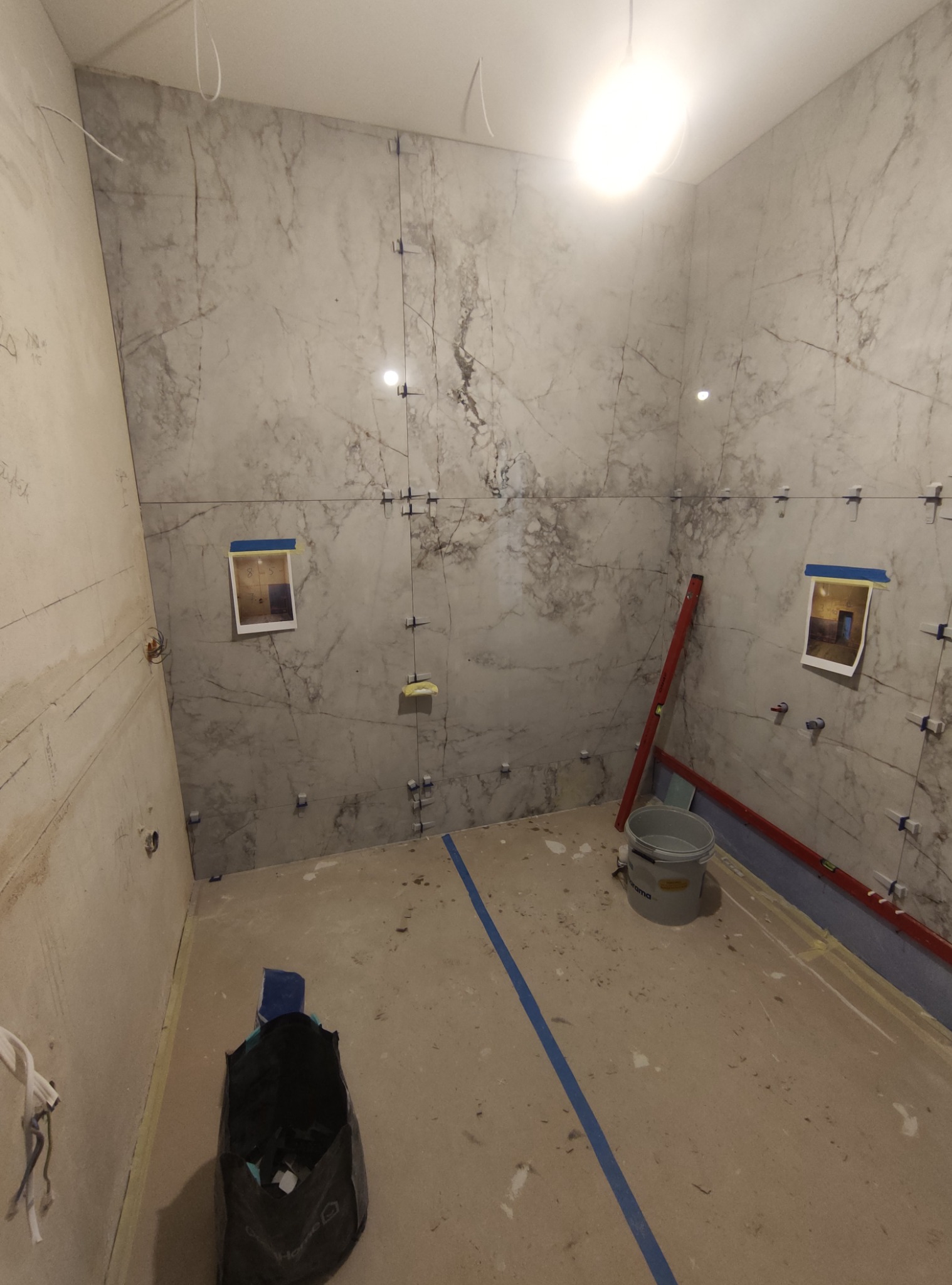 W trakcie układania marmurowych płytek w łazience, widoczne poziomice, krzyżyki dystansowe, przymocowane zdjęcia referencyjne na ścianie i wiadro z klejem.