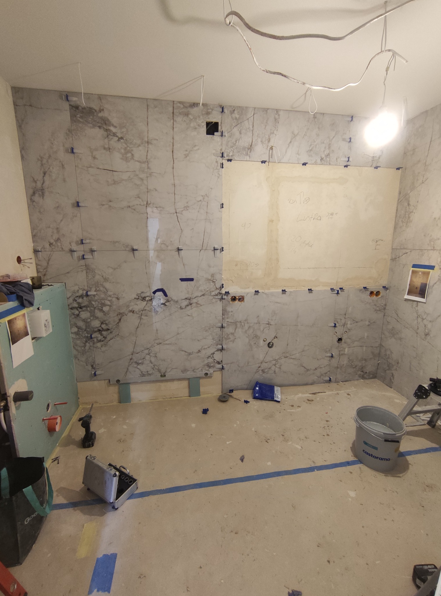 Łazienka w trakcie prac: ściany wyłożone płytkami imitującymi marmur, widoczne krzyżyki dystansowe, przygotowane punkty instalacyjne, notatki na ścianie, narzędzia i wiadro Castorama na podłodze.