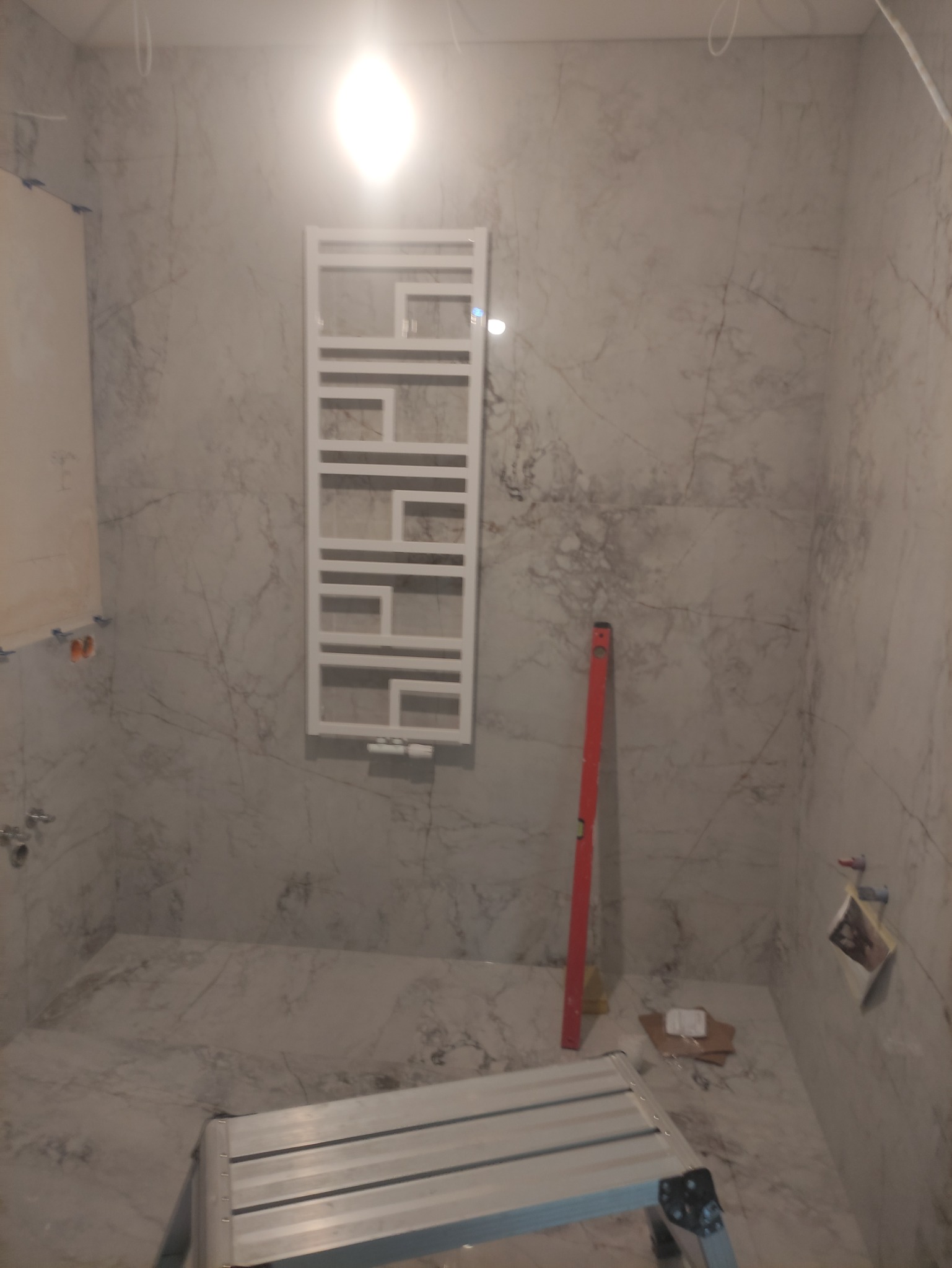 Łazienka w trakcie prac wykończeniowych: marmurowe płytki na ścianach i podłodze, zamontowany biały grzejnik drabinkowy, poziomica oparta o ścianę, drabina aluminiowa w pomieszczeniu.