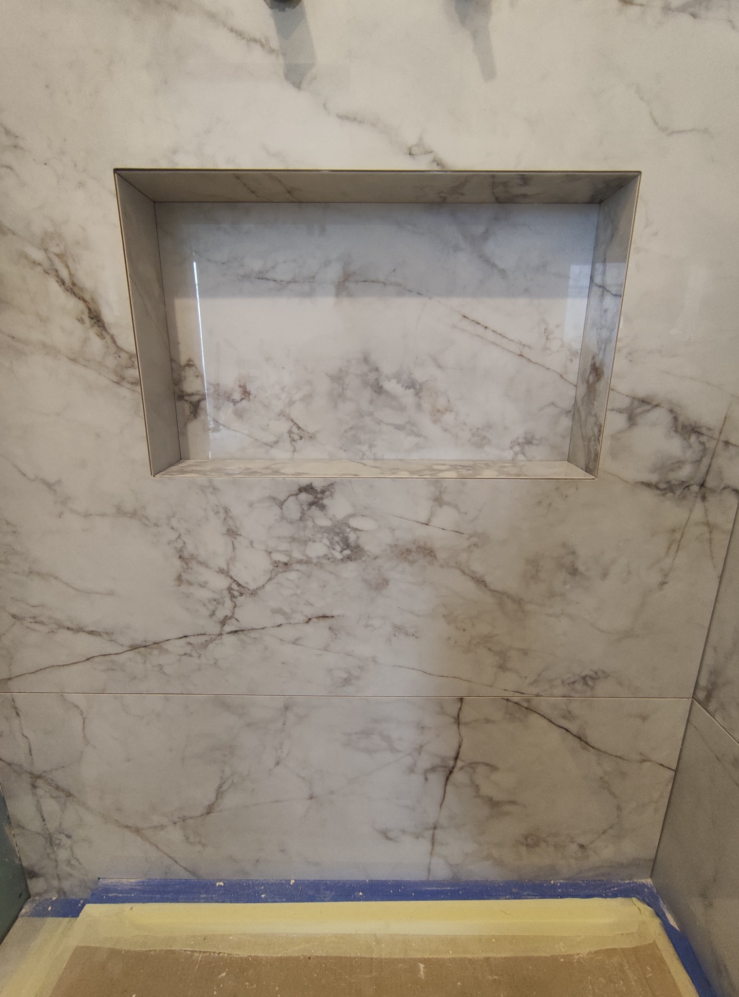 Wnęka prysznicowa wyłożona płytkami imitującymi marmur, z widocznym fragmentem podłogi zabezpieczonym taśmą malarską.