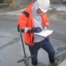 Geolog w pomarańczowej kurtce i kasku zapisuje dane z badania gruntu przy użyciu sondy, na placu budowy. W tle widoczne zabezpieczenia wykopu.