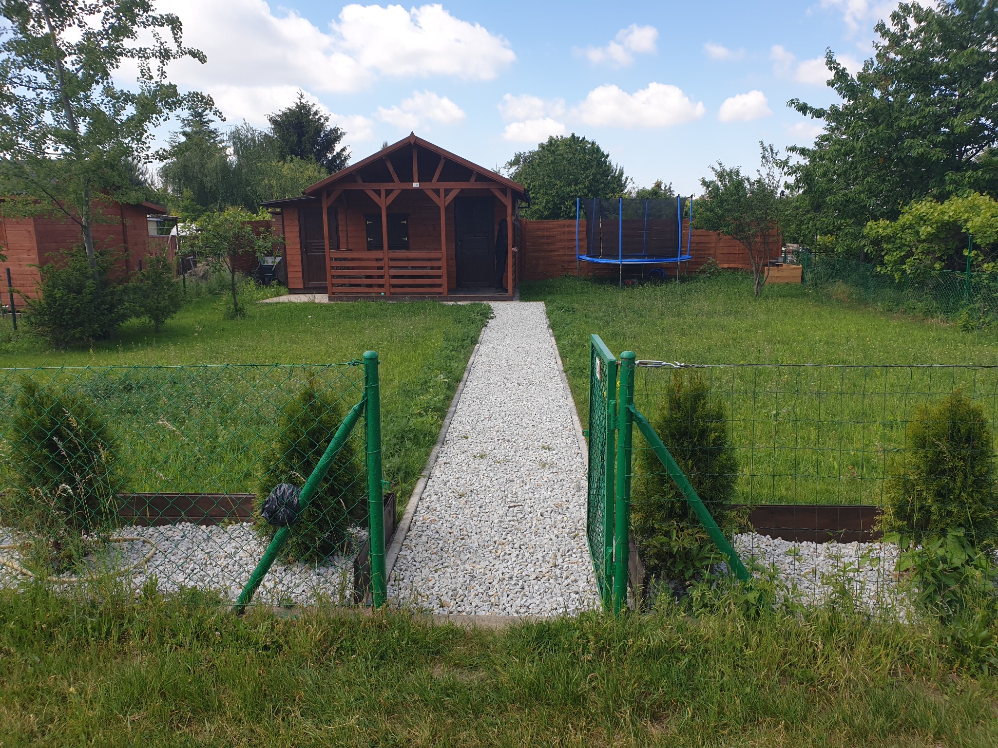 Widok na drewnianą altanę ogrodową z ciemnymi drzwiami, do której prowadzi ścieżka wysypana białym żwirem, widziana przez zielone ogrodzenie z otwartą furtką; w tle trawnik, trampolina i drzewa.
