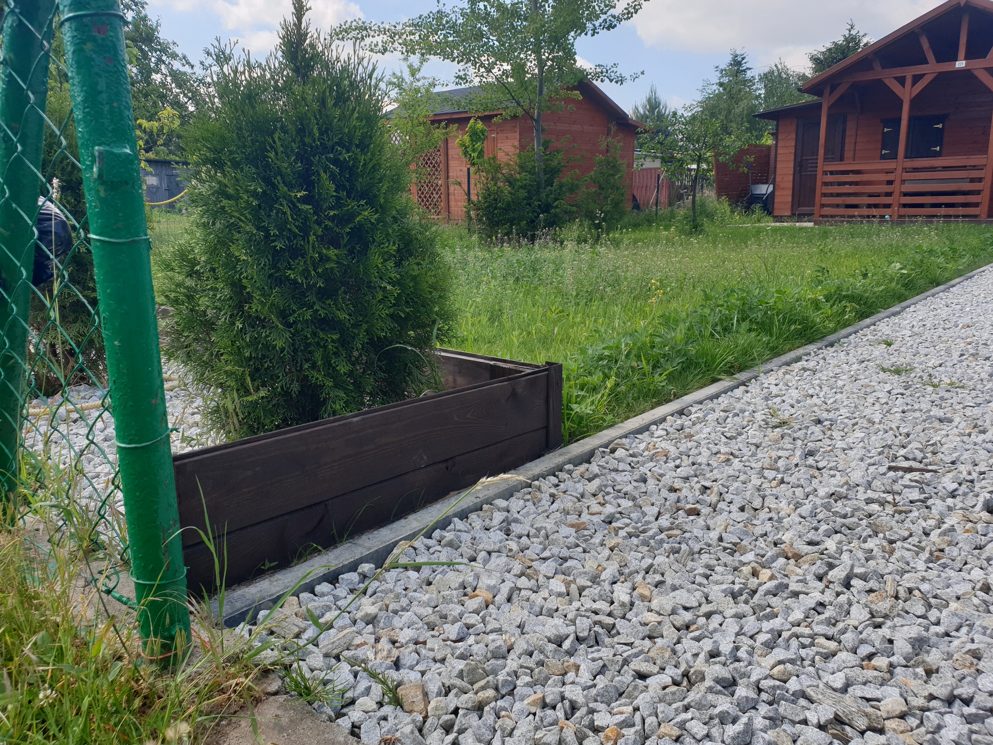 Widok na ścieżkę z drobnego kamienia prowadzącą do drewnianych domków letniskowych, z tujami w drewnianych donicach po lewej stronie i zielonym ogrodzeniem z siatki w pierwszym planie.