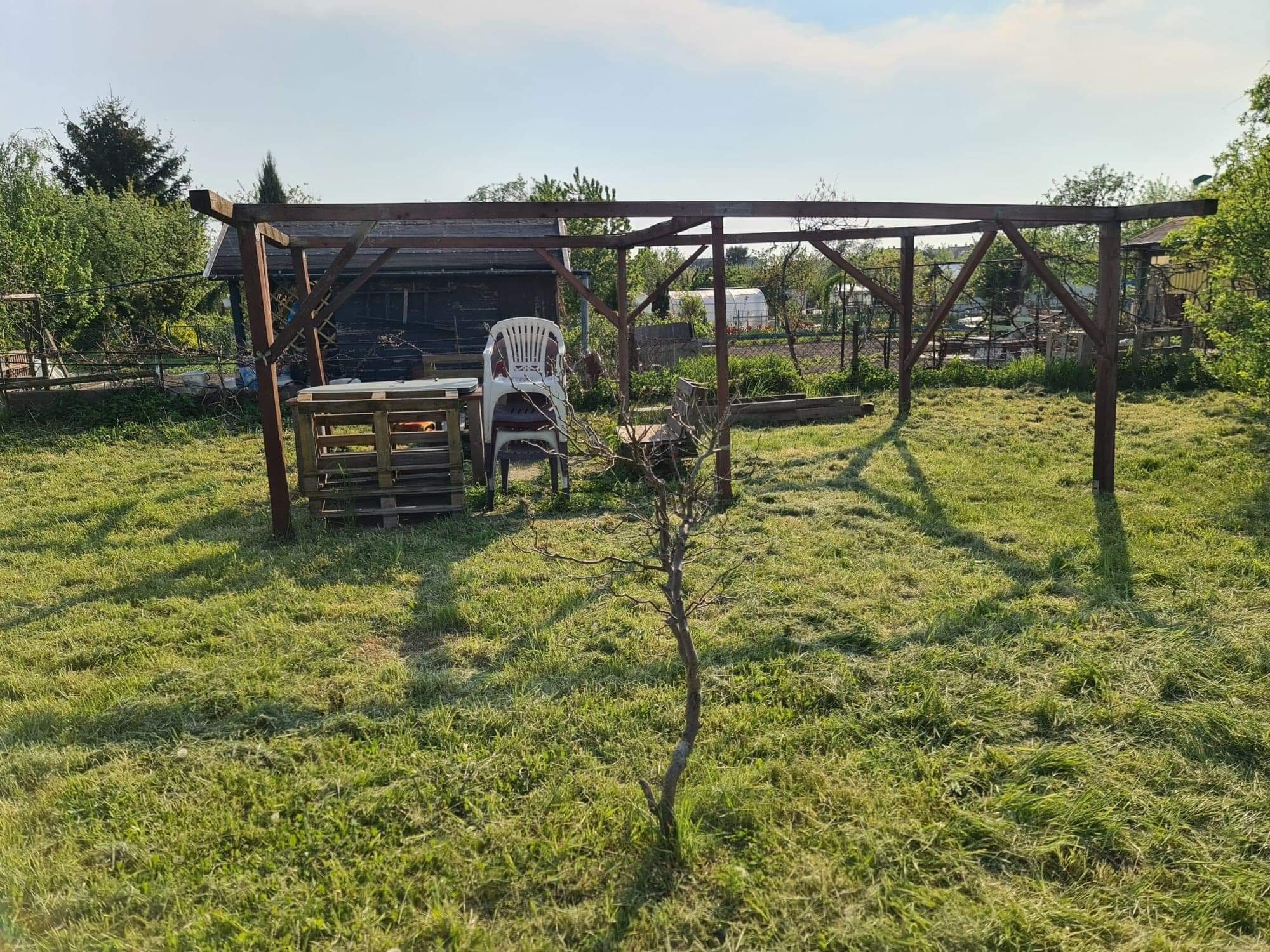 Niedokończona drewniana konstrukcja altany w ogrodzie, z widocznymi elementami konstrukcyjnymi, stosem palet i plastikowymi krzesłami w tle, na trawiastym terenie.