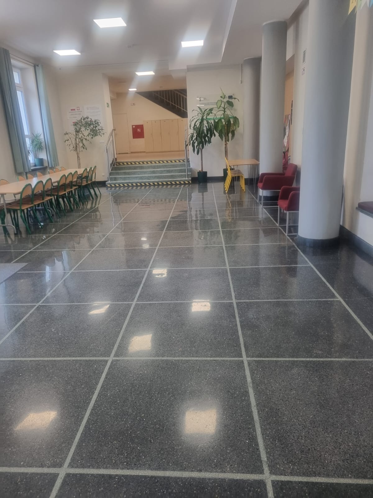 Błyszcząca, wypolerowana posadzka kamienna w holu budynku. Widoczne stoły, krzesła, kolumny i rośliny w donicach. Posadzka w ciemnych odcieniach szarości z jasnymi liniami podziału.