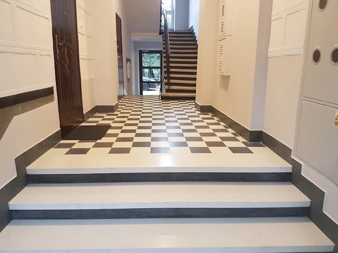 Klatka schodowa w kamienicy po remoncie: białe schody z szarymi krawędziami, podłoga w szachownicę, odnowione ściany i drzwi. Widok z dołu schodów.