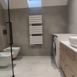 Jasna łazienka na poddaszu z oknem dachowym, szarymi płytkami, grzejnikiem drabinkowym, kabiną prysznicową typu walk-in z czarnymi profilami, umywalką nablatową i pralką w zabudowie.