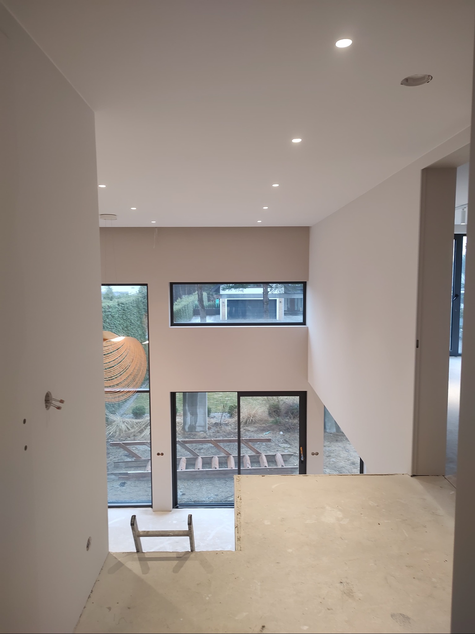 Wnętrze w trakcie wykańczania, widok z góry na betonową posadzkę, białe ściany, okna z widokiem na zewnątrz i aluminiową drabinkę.