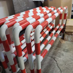 Stojaki ochronne, pomalowane w biało-czerwone pasy, ustawione rzędami w magazynie, gotowe do montażu.