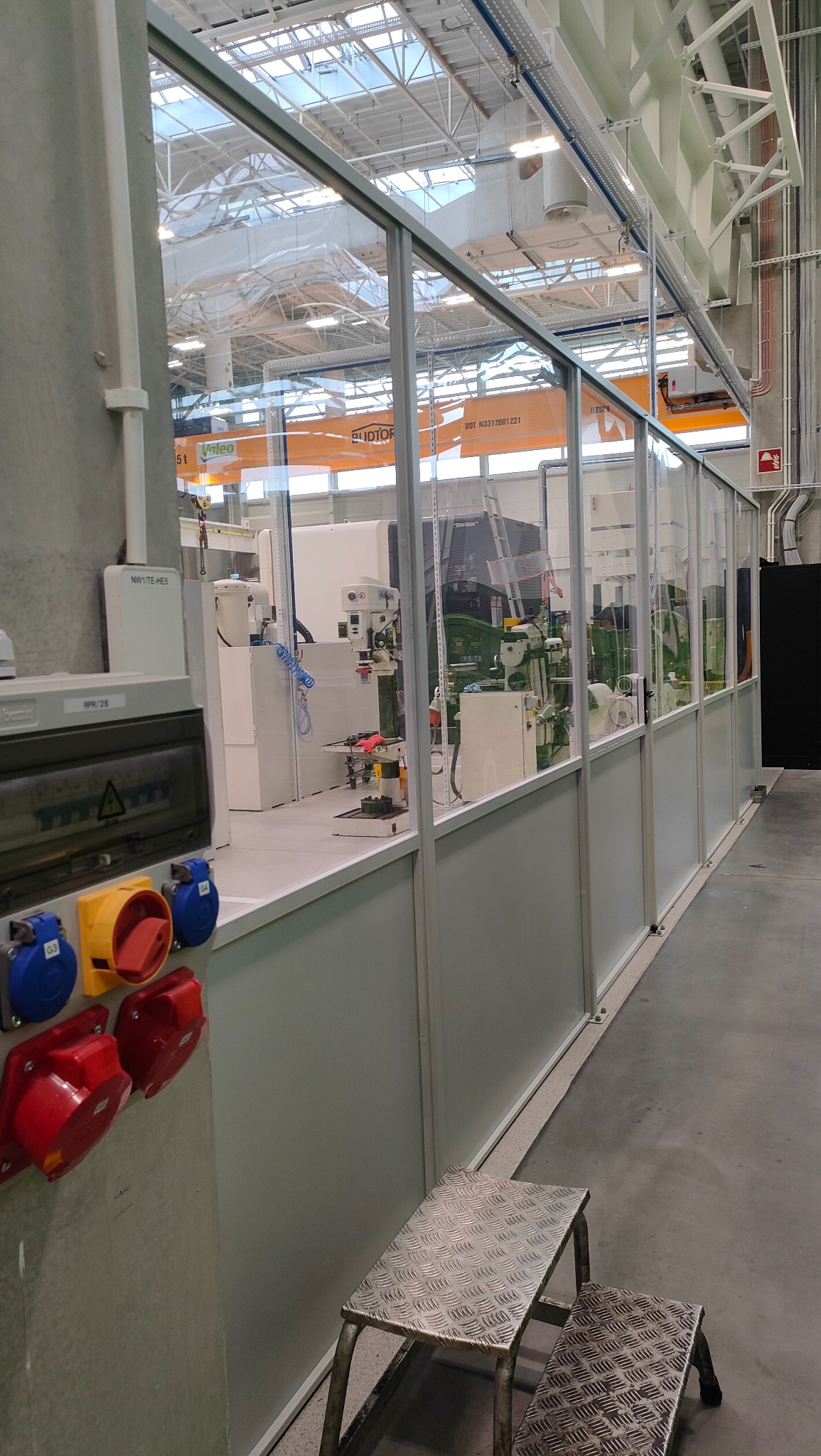 Przeszklona ścianka działowa w hali produkcyjnej, widoczne maszyny i urządzenia. Na pierwszym planie skrzynka elektryczna z gniazdami i metalowy podest.