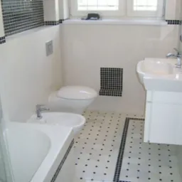 Nowoczesna łazienka z białymi płytkami i czarnymi akcentami mozaiki, widoczna umywalka na szafce, toaleta, bidet i wanna, całość utrzymana w minimalistycznym stylu.