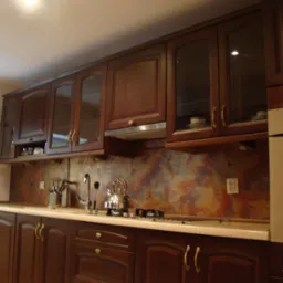 Klasyczna kuchnia z ciemnobrązowymi szafkami z przeszklonymi frontami, metalowym okapem i wielobarwną mozaiką na ścianie nad blatem.