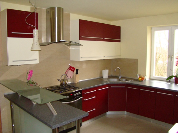 Nowoczesna kuchnia z bordowymi i kremowymi szafkami, stalowym okapem i barkiem z blatem ze szkła, oświetlona wiszącą lampą.