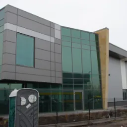 Nowoczesna hala z przeszkloną fasadą w Markach, z widoczną kabiną sanitarną i kontenerem na gruz, sugerującą trwające prace wykończeniowe.