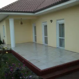 Wykończony taras z jasnych płyt, z obrzeżem z ciemnej kostki, przylegający do żółtej ściany domu z białymi oknami i kolumną, widok na rabatę kwiatową.