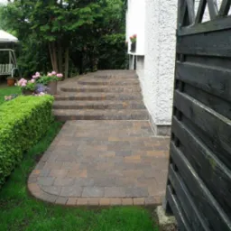 Brązowa kostka ułożona w ścieżkę i schody przed domem, obok przycięty żywopłot i czarny drewniany płot.