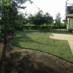 Nowo założony trawnik przylegający do domu z żółtą elewacją i kamiennym tarasem, widoczna świeżo przekopana ziemia pod nasadzenia wzdłuż trawnika.