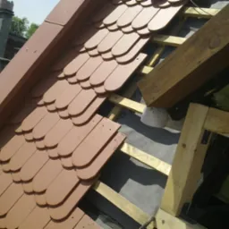 Montaż brązowej dachówki ceramicznej na drewnianej konstrukcji więźby dachowej, widoczne membrany dachowe i komin w tle.