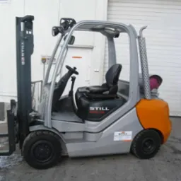 Szaro-pomarańczowy wózek widłowy STILL RX70-35 na zewnątrz, z metalową konstrukcją kabiny i czarnym siedzeniem. Widoczny z boku, na tle białej ściany budynku.