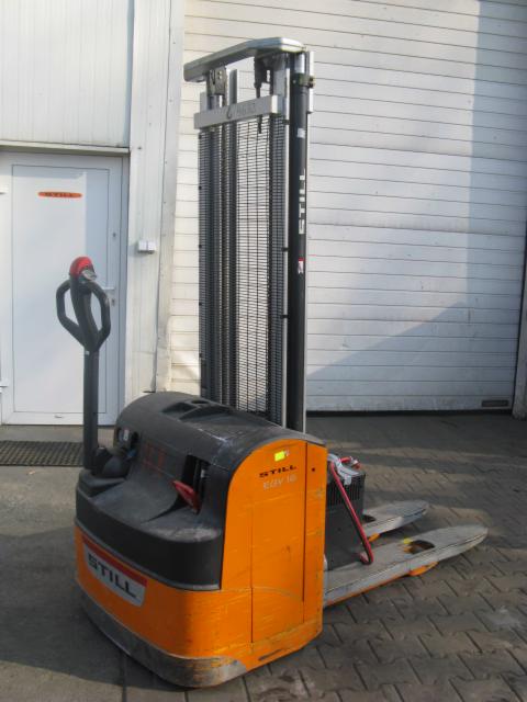 Pomarańczowo-czarny wózek paletowy elektryczny marki Still, model EGV 16, stoi na kostce brukowej przed szarą ścianą budynku z białą bramą.