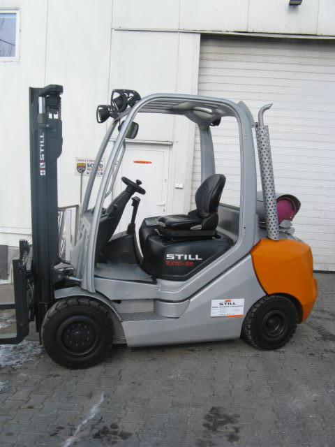 Szaro-pomarańczowy wózek widłowy STILL RX70-35 na zewnątrz, z metalową konstrukcją kabiny i czarnym siedzeniem. Widoczny z boku, na tle białej ściany budynku.