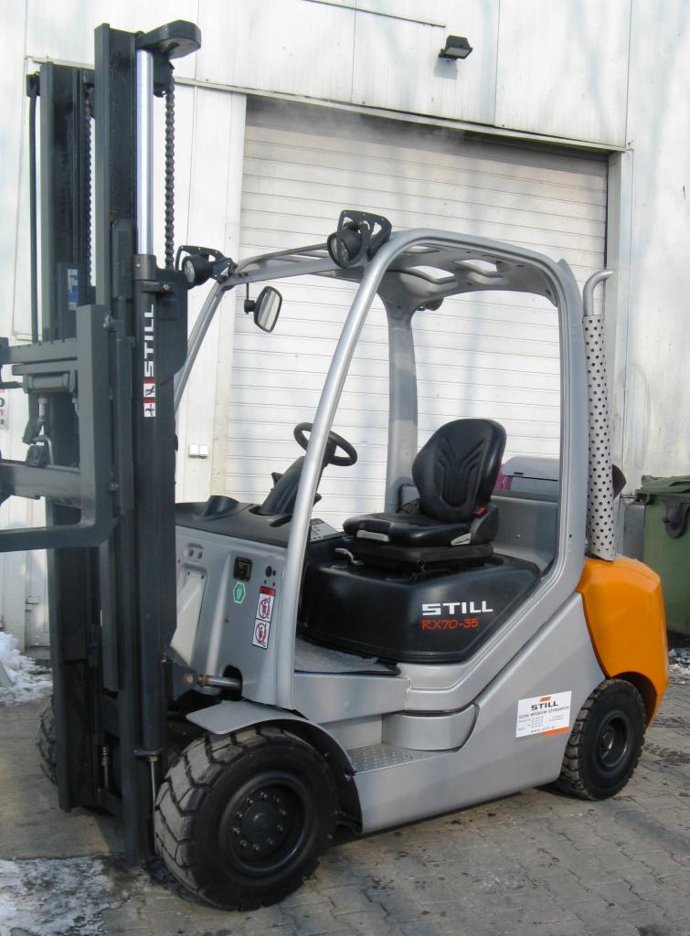 Szaro-pomarańczowy wózek widłowy Still RX70-35 z czarnym siedziskiem na tle białych drzwi magazynowych. Widoczny bok wózka i fragment wideł.