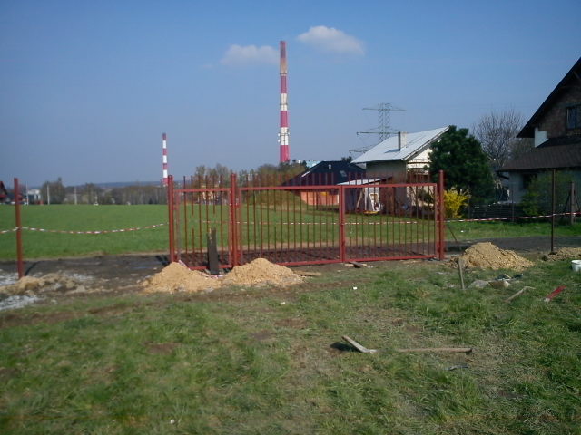 Montaż czerwonej, metalowej bramy wjazdowej na trawiastym terenie, w tle komin elektrowni i budynki mieszkalne. Widoczne narzędzia i usypany piasek.