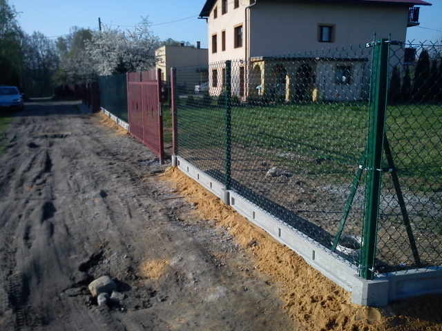 Nowo postawione ogrodzenie z siatki z podmurówką betonową i zielonymi słupkami na nierównym terenie, widoczna brama wjazdowa w kolorze bordowym i dom w tle.