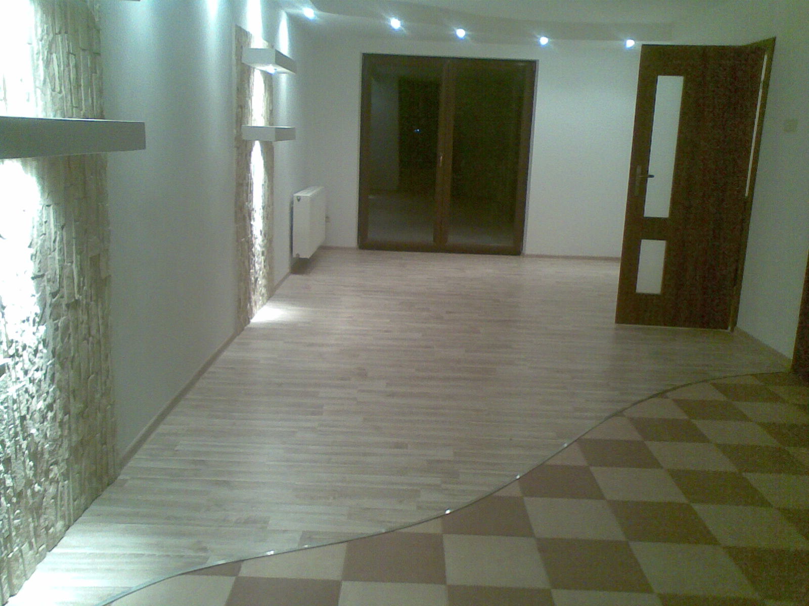 Wykończone wnętrze z dwiema rodzajami podłóg: imitacja drewna i szachownica, dekoracyjna ściana z kamienia z oświetleniem punktowym i półkami, oraz ciemne drzwi z przeszkleniami.