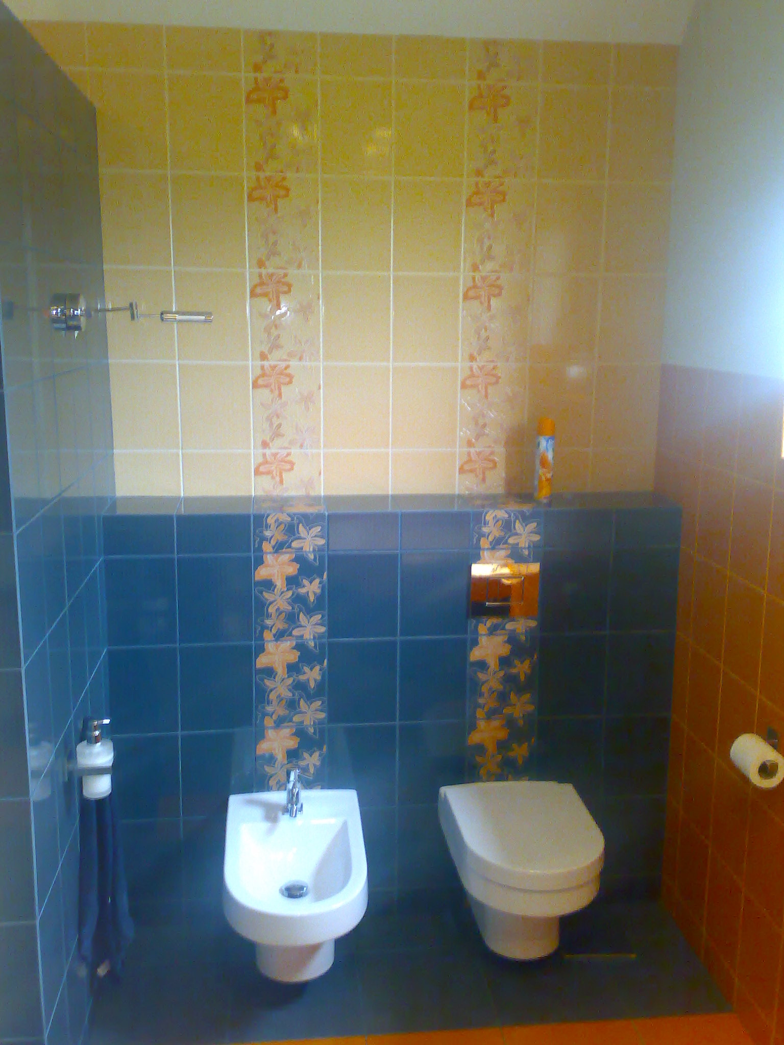 Wykończona łazienka z bidetem i toaletą w zabudowie, z niebieskimi i beżowymi płytkami z kwiatowym wzorem, ręcznikiem i dozownikiem mydła. Widoczny fragment prysznica.
