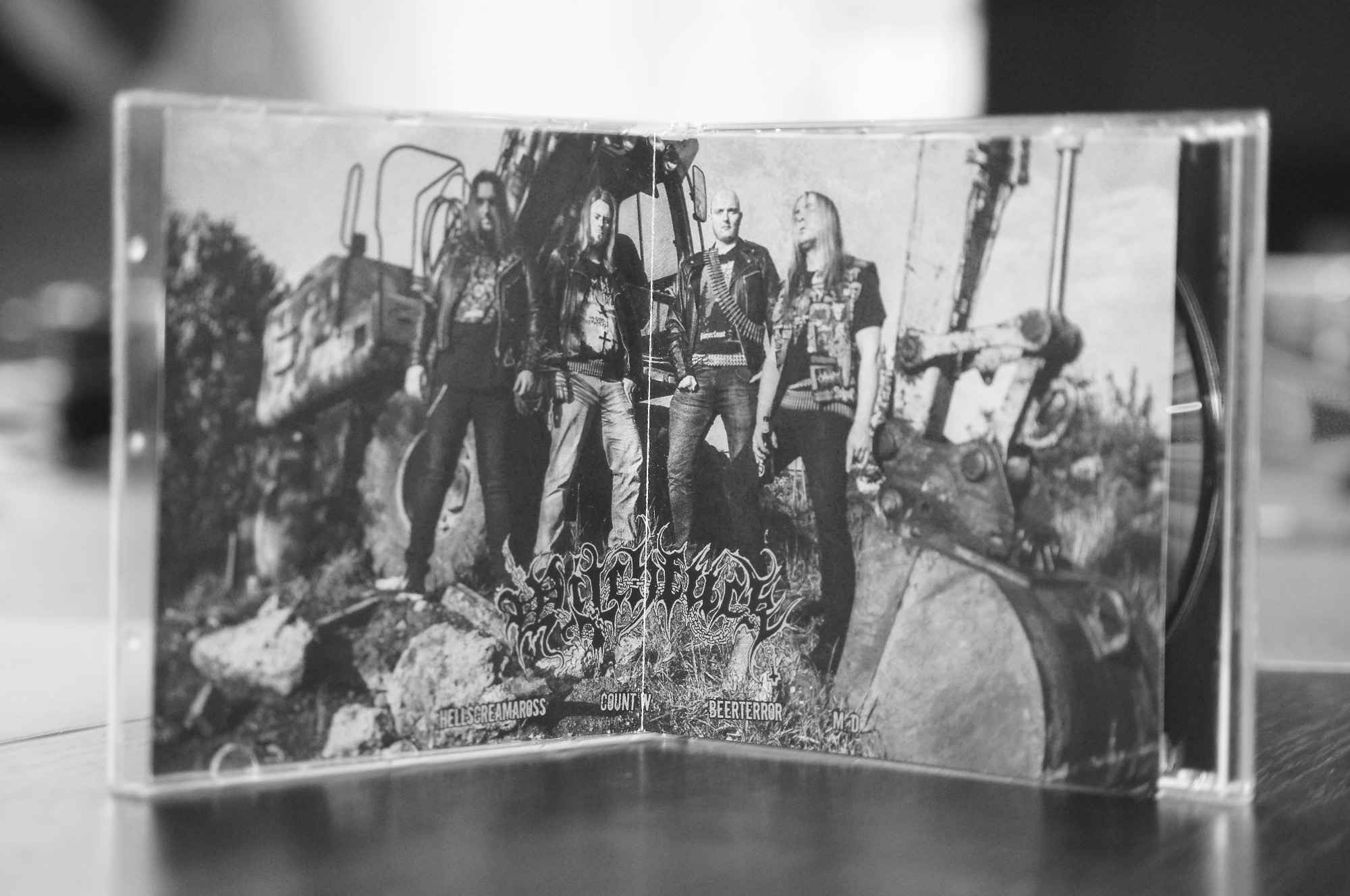 Czarno-białe zdjęcie okładki płyty zespołu metalowego z członkami zespołu na tle maszyny budowlanej, w plastikowym pudełku na drewnianym stole.