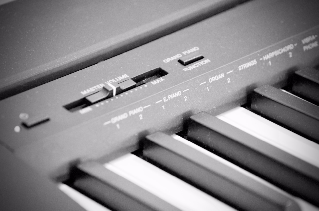 Czarno-białe zbliżenie na panel sterowania i klawiaturę pianina cyfrowego, z widocznymi suwakami głośności i przełącznikami funkcji, ustawionymi na różne brzmienia instrumentów.