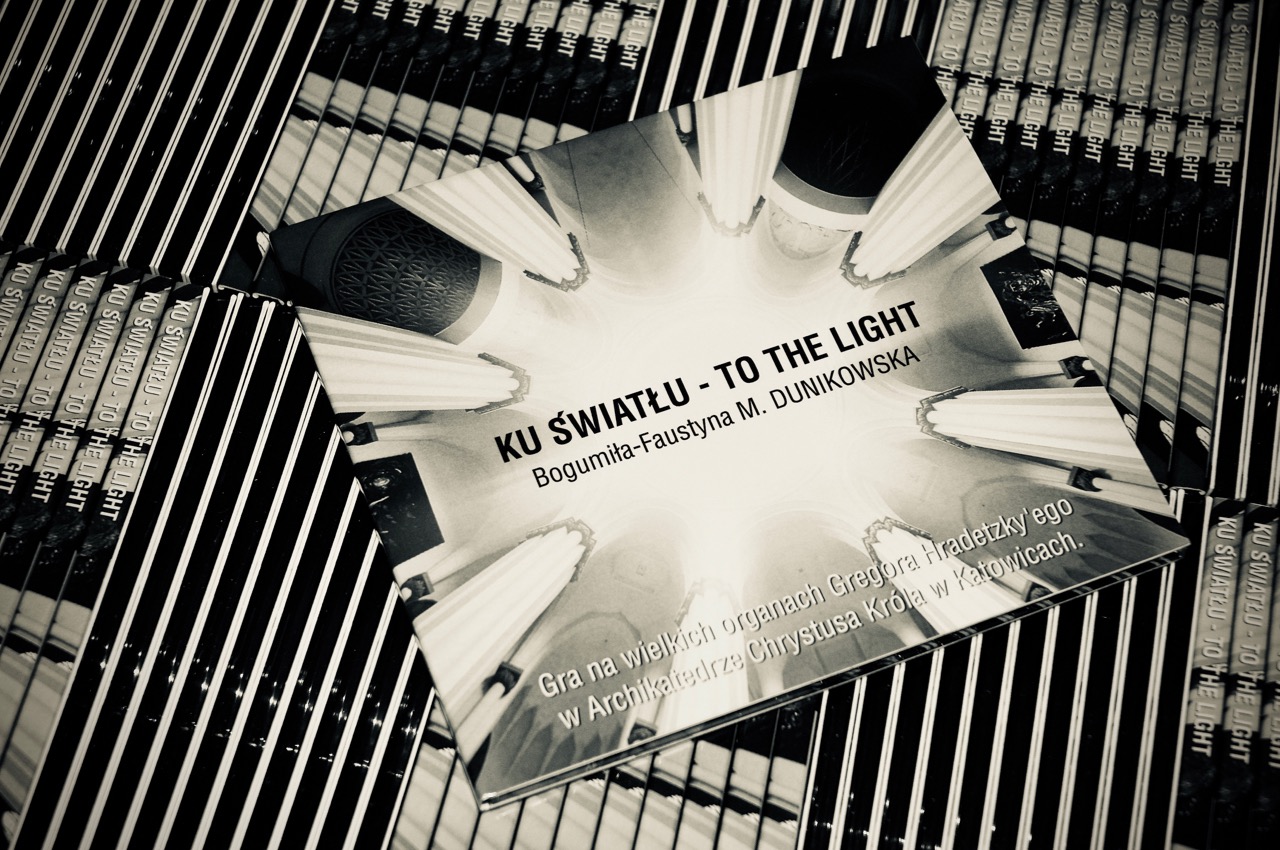 Czarno-białe zdjęcie okładki płyty CD 'Ku Światłu - To the Light' Bogumiły-Faustyny M. Dunikowskiej, z motywem architektonicznym wnętrza katedry, ułożonej na tle innych egzemplarzy płyty.