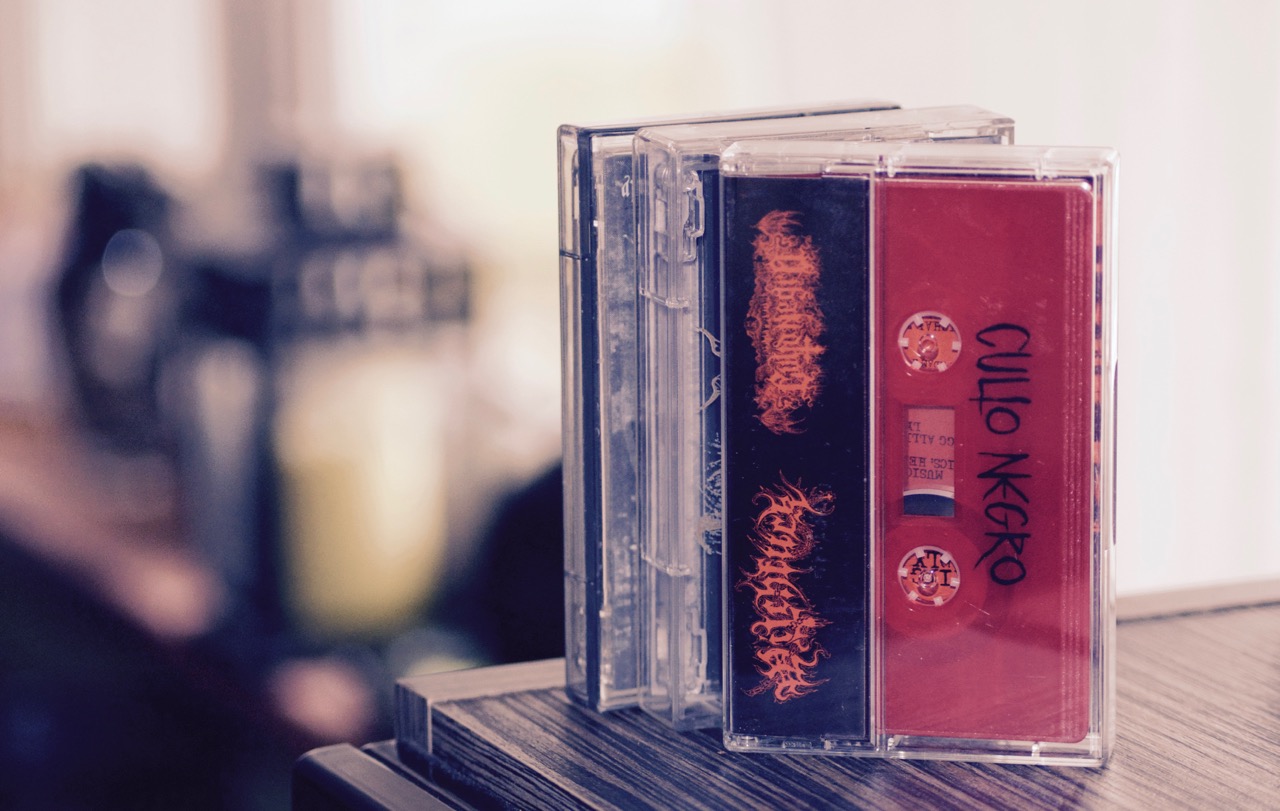 Cztery kasety magnetofonowe w przezroczystych pudełkach ustawione jedna za drugą, z czego dwie z widocznymi okładkami w kolorach czarnym i czerwonym, na drewnianej powierzchni. Okładki kaset...