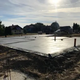 Wylany, świeży fundament płyty na gruncie, zabezpieczony czarną folią, widoczne wystające rury i pręty zbrojeniowe, w tle zabudowa jednorodzinna i błękitne niebo z chmurami.
