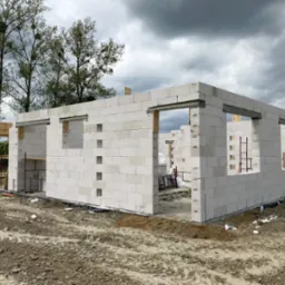 Wznoszenie ścian parterowego domu z białych bloczków, widoczne otwory okienne, drewniane szalunki i rusztowania wewnątrz, błotniste podłoże, pochmurne niebo w tle.