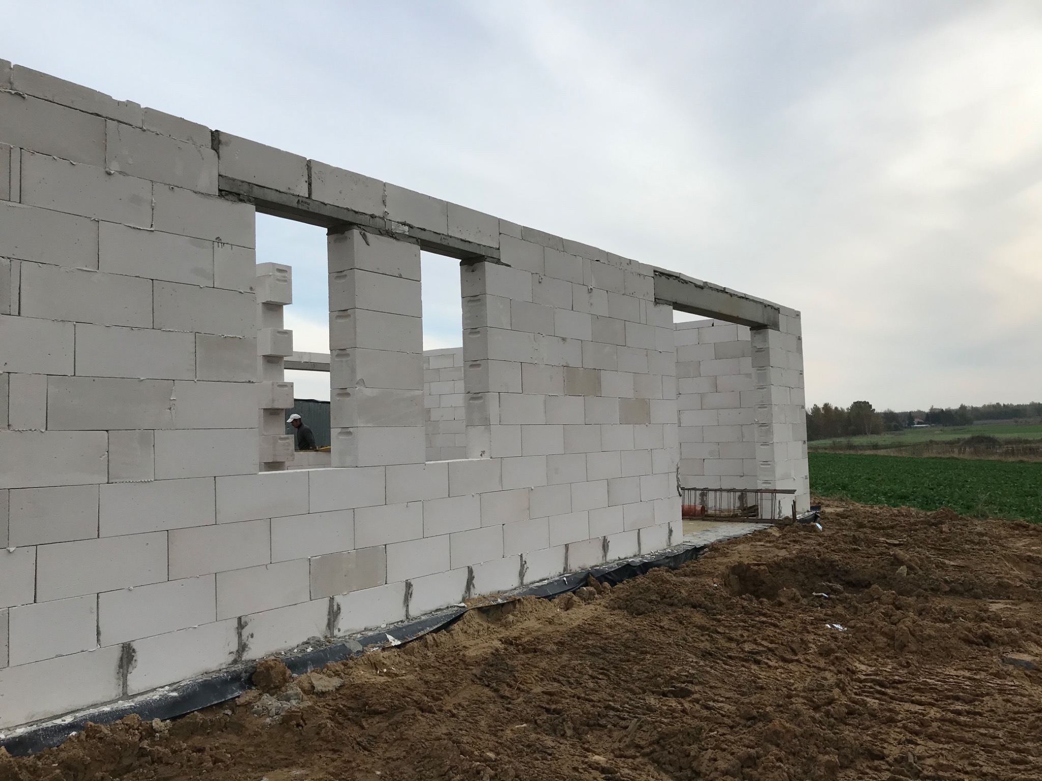 Wznoszenie ścian domu z białych bloczków z widocznymi otworami okiennymi i nadprożami, w tle krajobraz polny i pochmurne niebo.