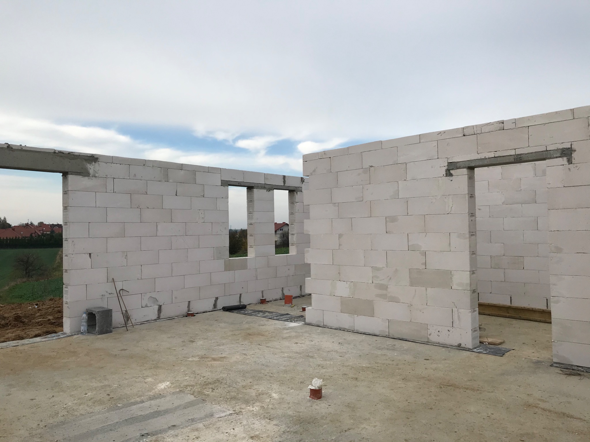 Wnętrze surowego budynku z białych bloczków, widoczne otwory okienne i drzwiowe ze stalowymi nadprożami, na zewnątrz widok na zielone pola i zabudowania.