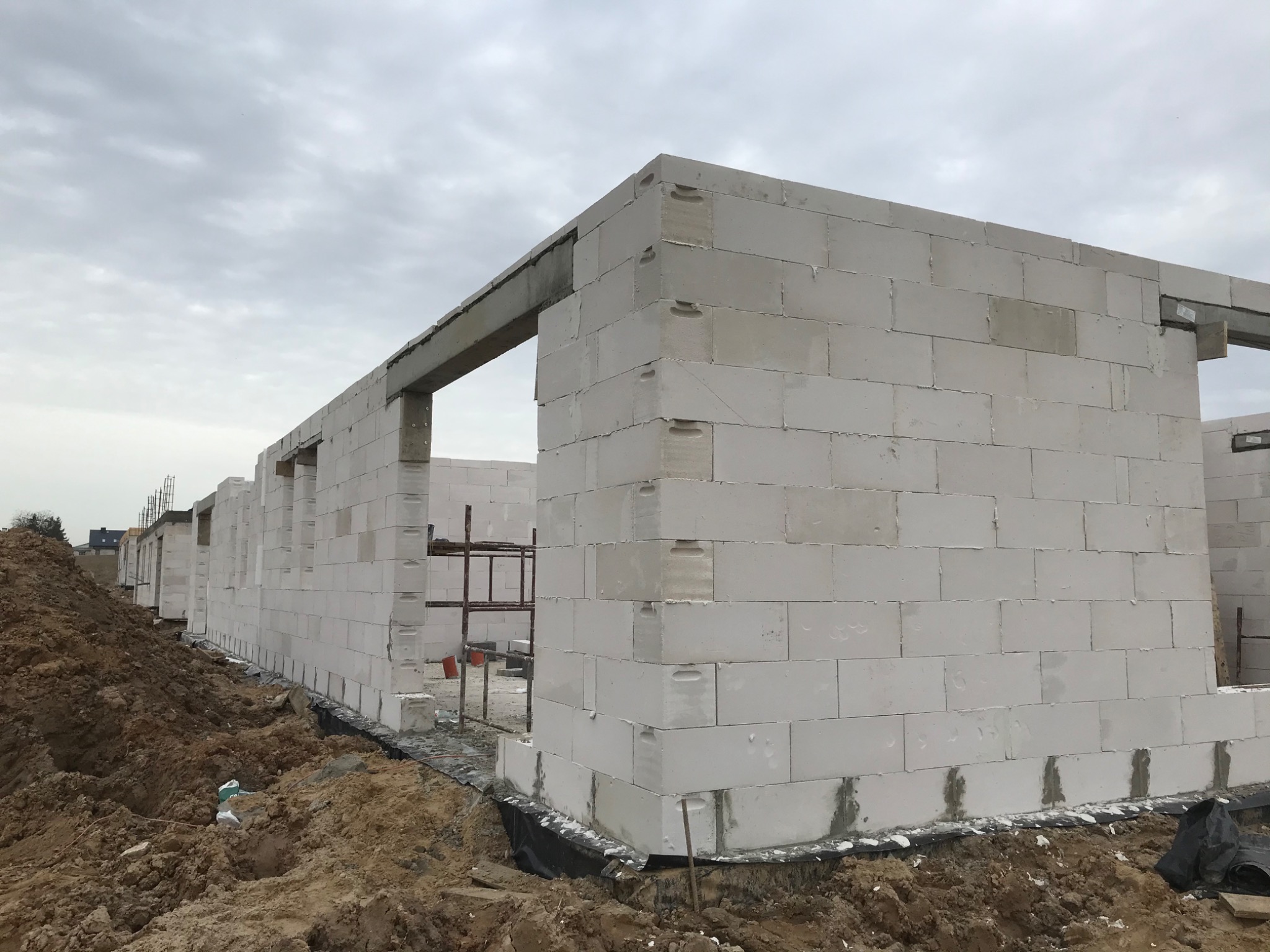 Wznoszenie ścian parterowego domu z betonu komórkowego na fundamencie, widoczne zbrojenie nad otworami okiennymi, obok hałda ziemi.