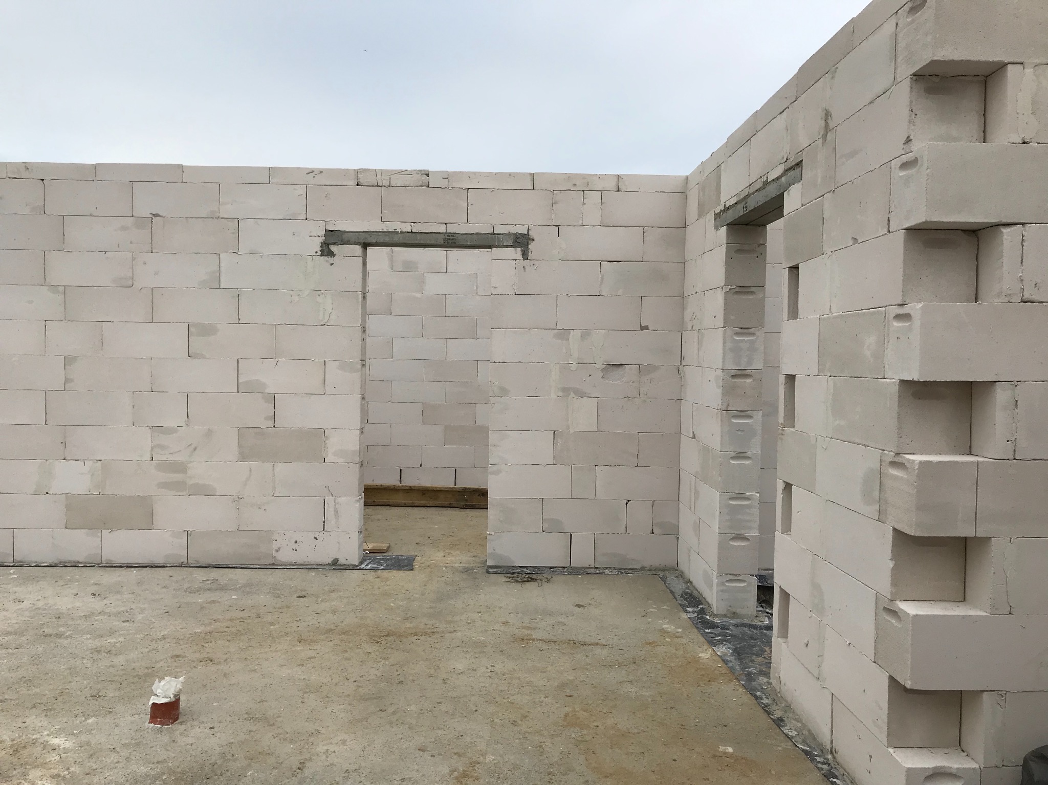 Wnętrze w trakcie budowy z białych bloczków, widoczne otwory drzwiowe z nadprożami, surowy beton na podłodze i fragment muru z przesuniętymi bloczkami.